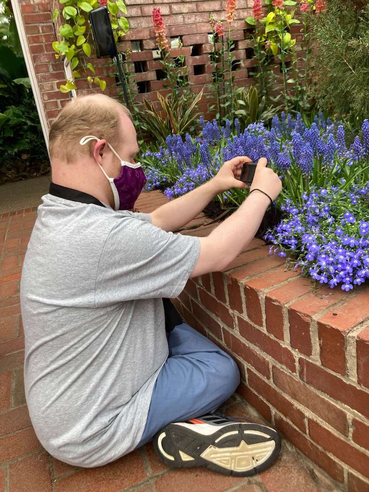 Tony sitting cross-legged taking a photo of purple flowers in a brick-lined flowerbed at Phipps Conservatory