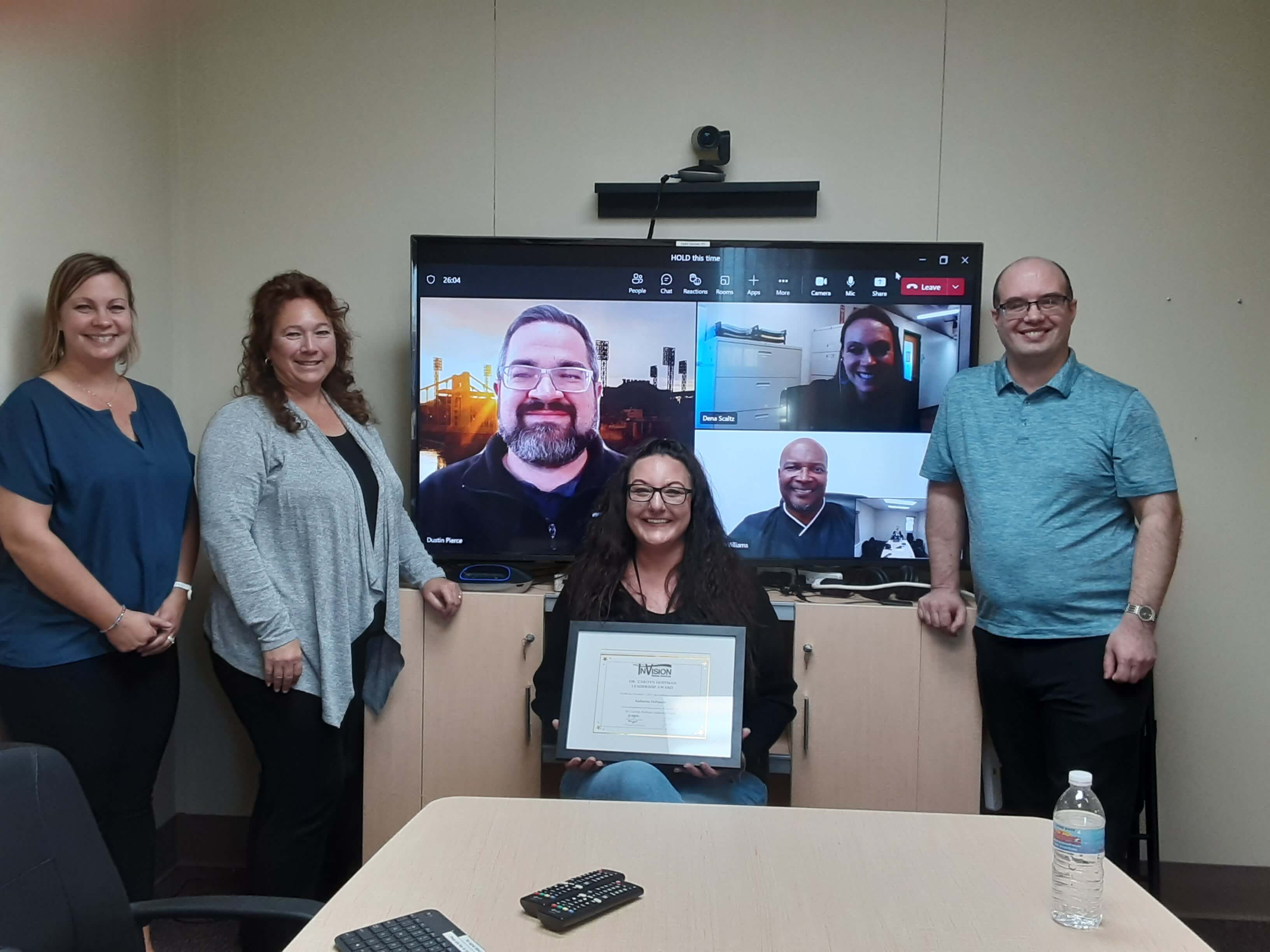 Katherine DeNunzio, seated, holds her leadership award in front of a TV screen with InVision leadership on it and flanked by additional InVision leadership